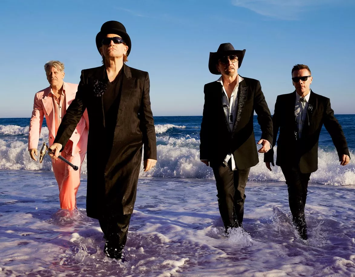 U2 üyelerinin yer aldığı bir fotoğrafın ön planında Carnaval de Luz albümünü çağrıştıran sıcak ve festival hissi veren ışıklar, arka planda sahne atmosferi ve Dolly Parton'a ince bir gönderme.